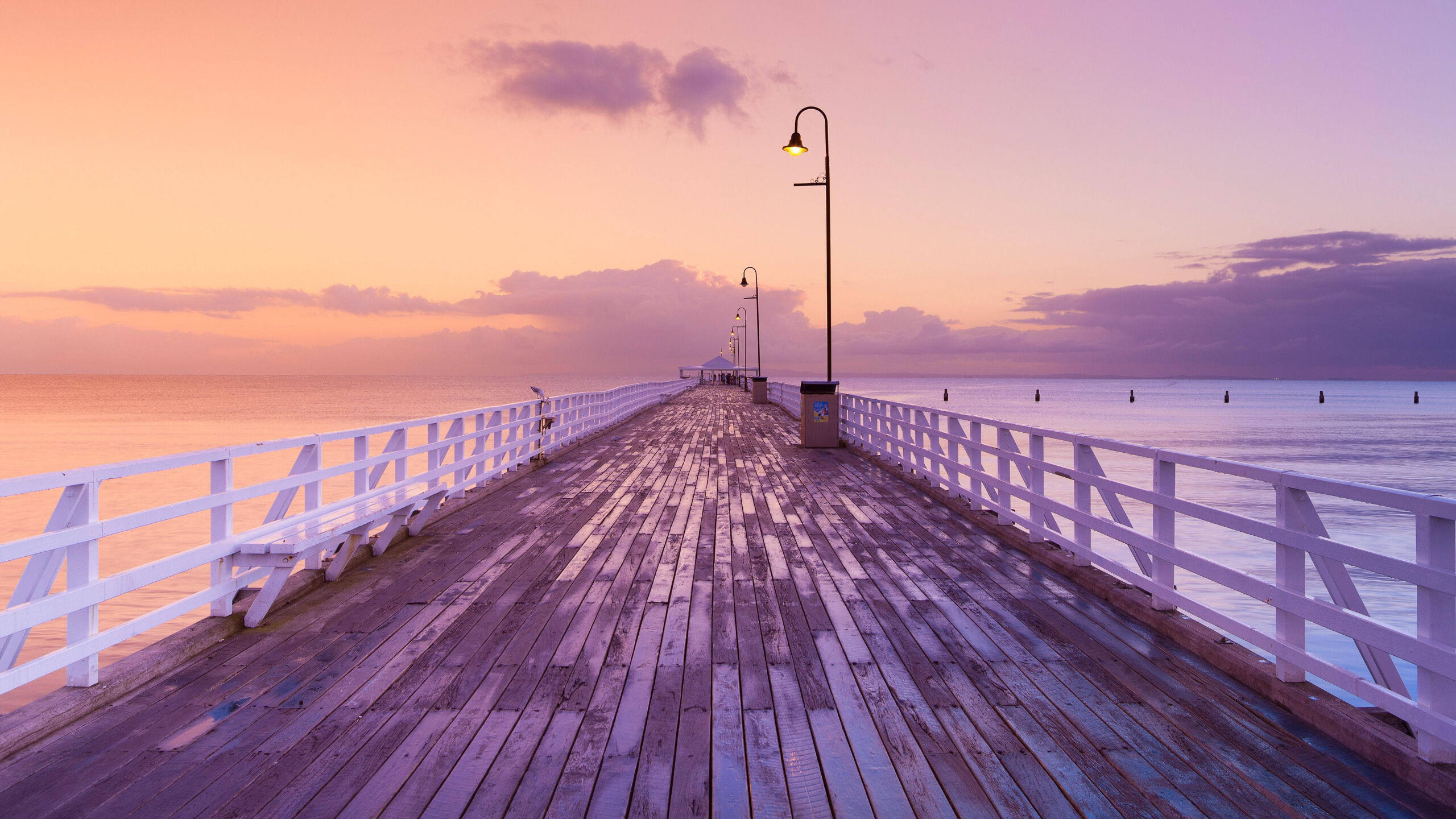 A wooden pier is pictured. On one side, the sky is orange. On the other side, the sky is purple. It is sunset. We are looking down the pier.