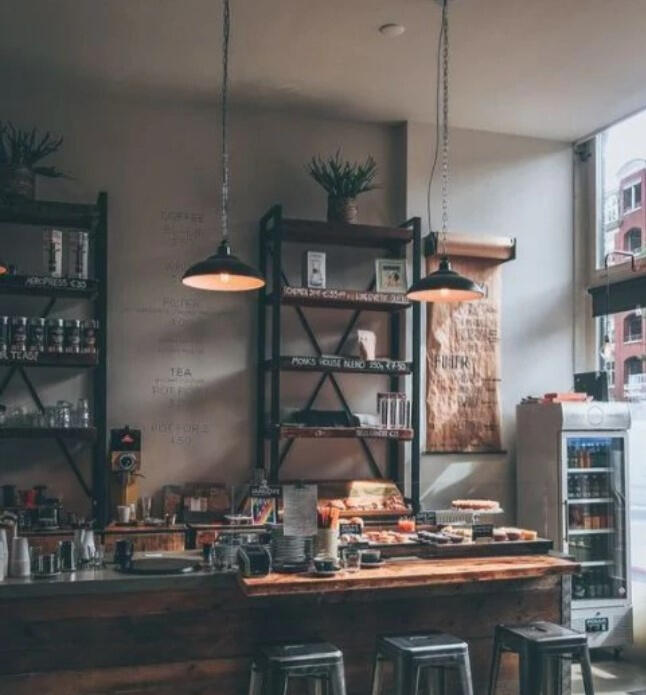 An industrial cafe is pictured. Light is coming from a window on the right side. There are 2 lights hanging over a bar area with shelves in the background.