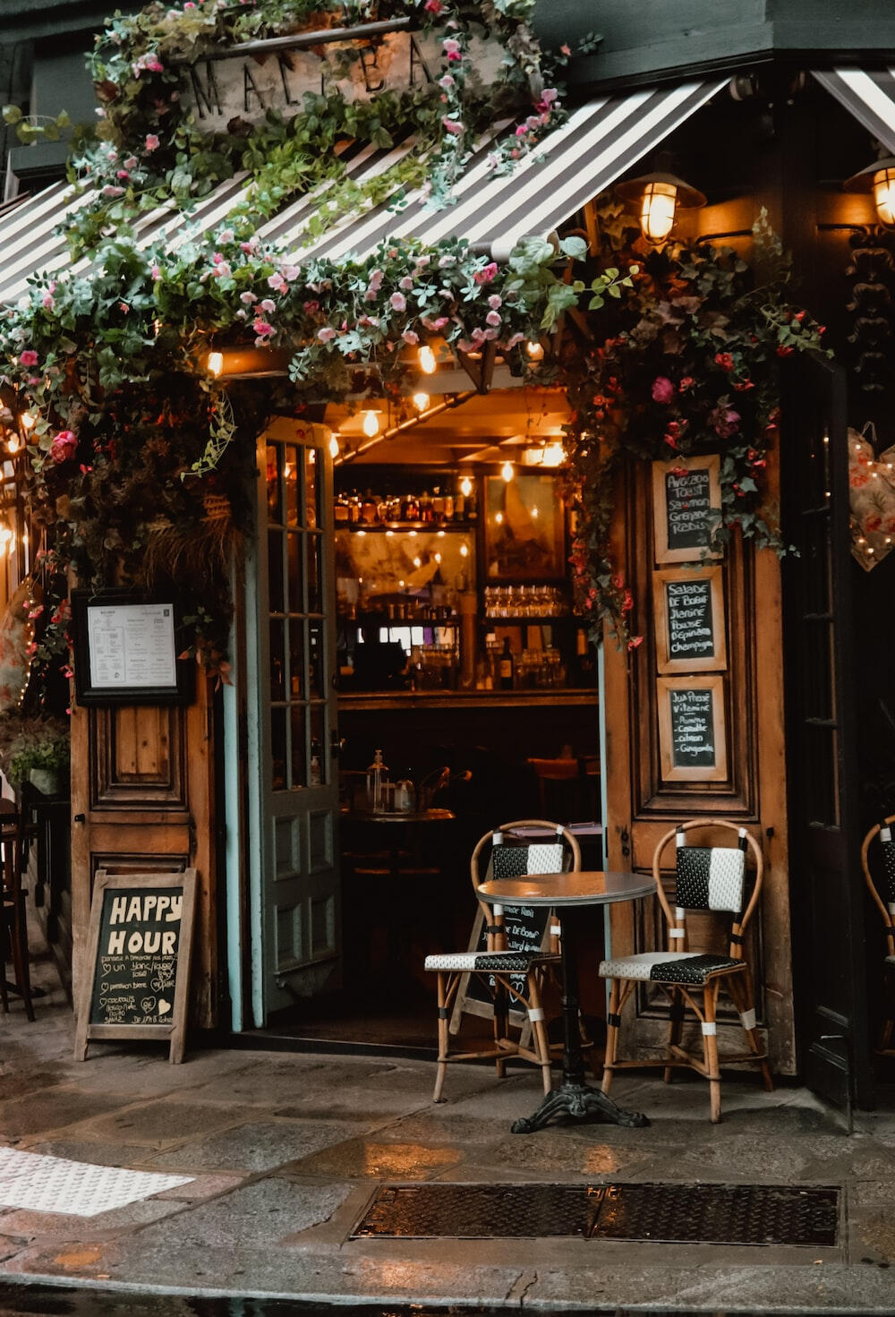 A view of the entrance to a cafe is pictured. There are flowering vines surrounding the canopy roof. There is a table with 2 chairs outside. It has recently rained.